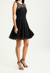 Černé šaty s přiléhavým korzetem a rozšířenou sukní. Obsahují vyšívané detaily na výstřihu. S párem černých sandálů na podpatku.