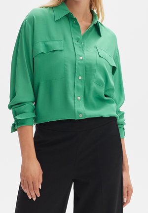 Femme portant une chemise verte à manches longues avec poches à rabat et un pantalon noir, debout devant un fond uni.