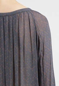 Průhledná blůza se nařasenými rameny, s tmavě modrou základnou a malým geometrickým vzorem v měděné a modré. Ideální pro vrstvené outfity.