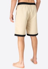 Beige swim shorts featuring a black elastic waistband and hem, with a side pocket and minimal detailing. Soft, lightweight fabric.
