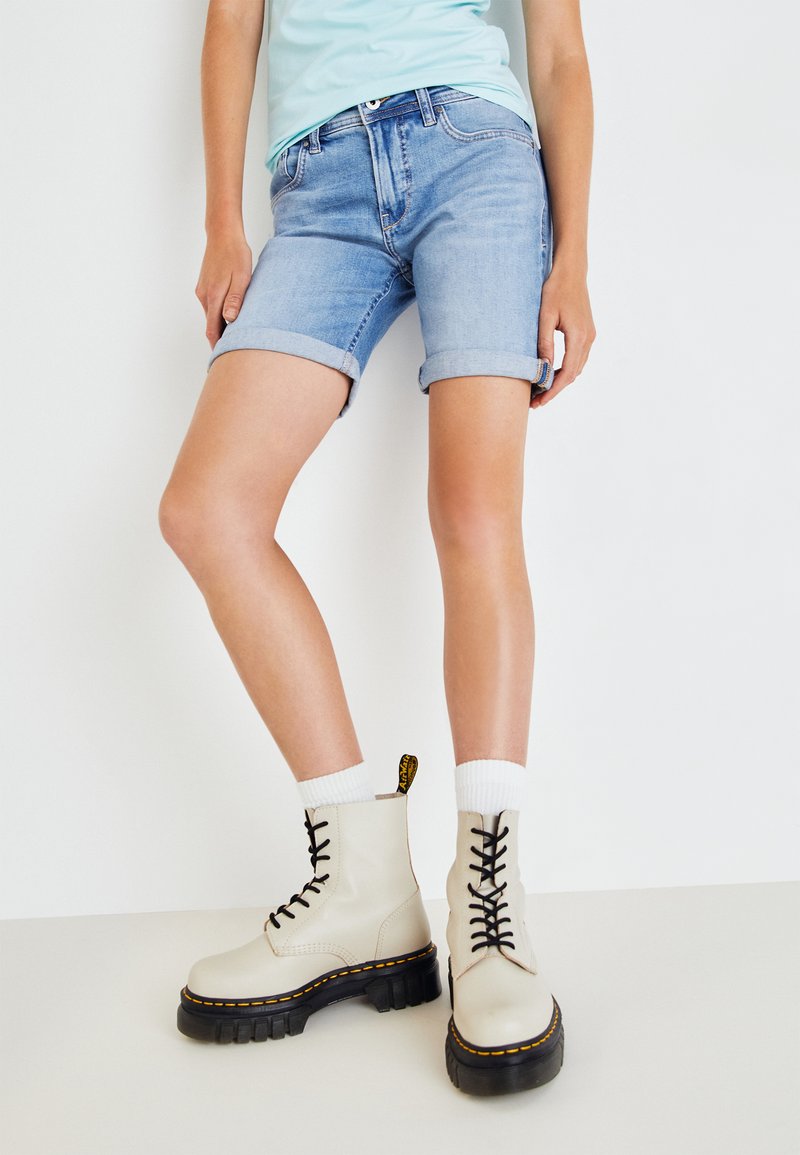 Šviesiai mėlyni džinsiniai šortai su apvyniotais kraštais, derinami su smėlio spalvos nėriniais palenkiamais ilgais batais, turinčiais masyvų juodą padą ir geltonus siūlus.