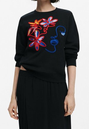 Sweatshirt noir orné de fleurs colorées brodées en rouge, bleu et jaune, avec un col rond et des poignets côtelés. Associé à une jupe plissée noire.