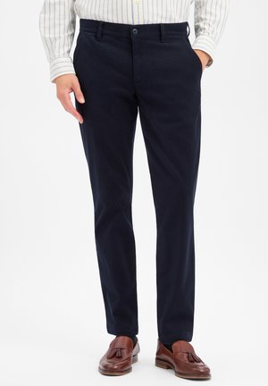 Man draagt marineblauwe broek, bruine tasselloafers en een wit overhemd met dunne blauwe verticale strepen. Eén hand in de zak.