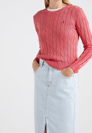 Femme portant un pull rose corail à torsades avec un sous-pull blanc et une jupe en jean bleu clair taille haute avec une fente à l'avant.