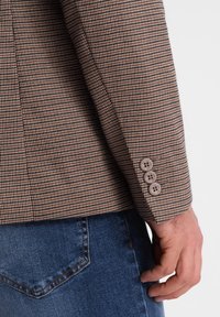 Brązowa marynarka w kratę w jodełkę z trzema brązowymi guzikami na mankiecie, połączona z niebieskimi dżinsami. Widoczny detal faktury tkaniny.