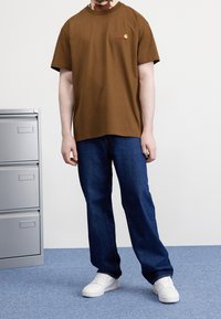 Camiseta de algodón marrón con cuello redondo y mangas cortas, que presenta un pequeño logotipo bordado. Combinada con jeans azul oscuro y zapatillas blancas.