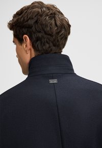 Marineblauwe wollen jas met hoge kraag, voorzien van een klein merklabel aan de achterkant. Textuur lijkt glad en solide.