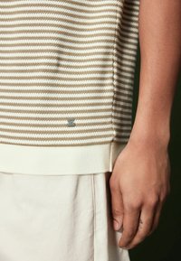 Pull en maille rayé beige et crème, avec col et ourlet côtelés. Petit détail logo près du bas. Texture lisse.