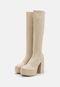 Botas altas beige de tacón, hechas de cuero suave, con una plataforma rectangular y tacones gruesos. Detalles de costura visibles en la parte superior.