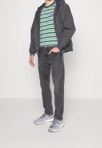 Grå denimjacka, randig grön och lila t-shirt, grå jeans och grå sneakers med vita detaljer, stående mot en vit bakgrund.