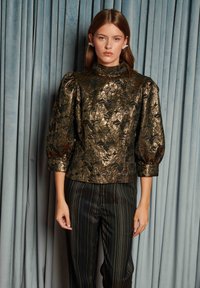 Young woman wears metallic gold floral blouse with puffed sleeves and black pinstriped trousers, standing against blue velvet curtains.