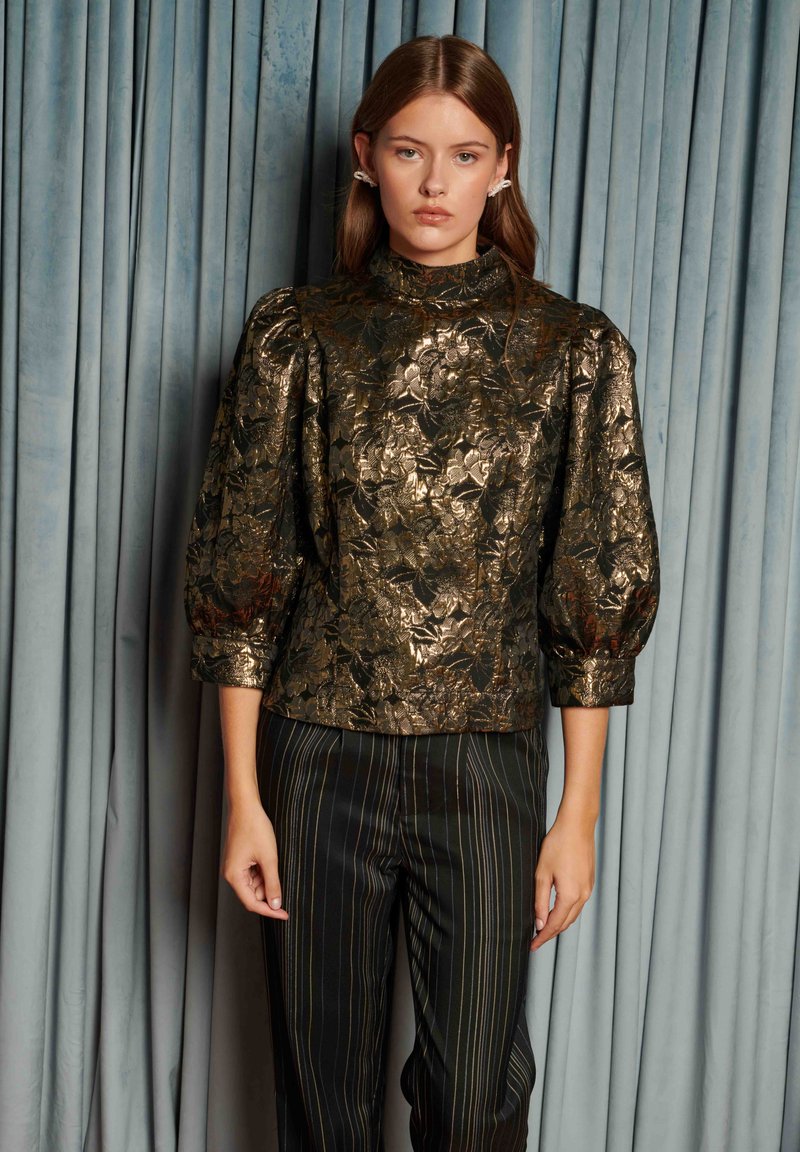Young woman wears metallic gold floral blouse with puffed sleeves and black pinstriped trousers, standing against blue velvet curtains.