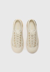 Beige canvas sneakers met gouden veterogen, vetersluiting en subtiele patronen. Ronde neus en een getextureerde rubberen zool. Merkenlogo zichtbaar.