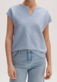 Světle modrý krátký rukáv top s výstřihem do V, s texturovanými horizontálními pruhy. Nosič oblečený s světle modrými džíny, které ukazují neformální styl.