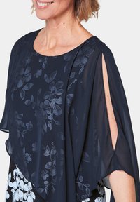 Marineblaue Bluse mit floralem Muster, durchscheinendem Stoff, weitem Ausschnitt und kurzen Ärmeln mit Schlitzen für zusätzlichen Detail.