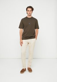 Brun kortermet t-skjorte med blått logo, kombinert med lysebeige bukser og brune semskede slip-on sko. Minimalistisk design, avslappet passform.