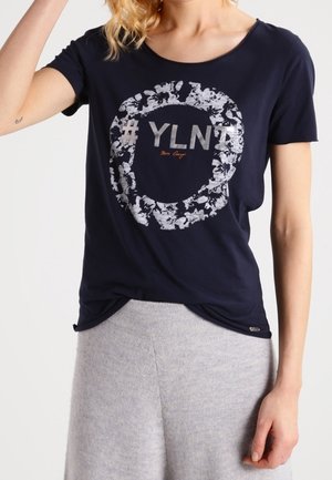 Osoba nosząca granatowy t-shirt z białym kwiecistym kołem i napisem „#YLN”, w zestawieniu z jasnoszarymi spodniami.