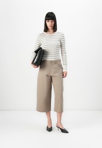 Vrouw in beige wijdvallende cropped broek, gestreept shirt met lange mouwen, zwarte puntige hakken, terwijl ze een grote zwarte clutch onder één arm houdt.