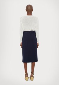 Pull blanc à manches longues associé à une jupe midi bleu marine et des sandales à talons beige, vu de l'arrière. Texture douce et lignes épurées.