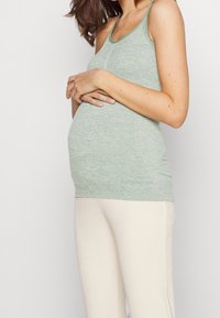Mint green tank top with thin straps, made of a soft, textured fabric. Fitted design complements a pregnant belly, paired with light, ribbed pants.