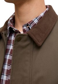 Chaqueta con cuello de pana marrón y tela lisa de color verde oliva, combinada con una camisa de franela a cuadros marrón rojizo y blanco.