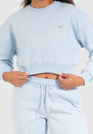 Vrouw draagt een lichtblauwe cropped sweatshirt en bijpassende joggingbroek, handen houden de zoom van de sweatshirt vast, waarbij de buik zichtbaar is.