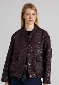 Donna che indossa una giacca viola scuro con paillettes, dotata di bottoni e tasche a pattina, abbinata a jeans blu scuro su uno sfondo grigio uniforme.