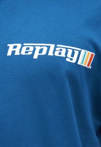 Μπλε ύφασμα με λευκό λογότυπο κειμένου "Replay" και τρεις κάθετες λωρίδες σε τιρκουάζ, πορτοκαλί και κόκκινο στα δεξιά του κειμένου.