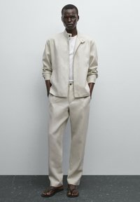 Helle beige Jacke mit Stehkragen, passende Hose und weißem T-Shirt. Trägt dazu braune Flip-Flops mit einer glatten Stoffoberfläche.