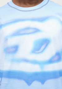 Άτομο που φοράει φωτεινό μπλε πουκάμισο με ένα θολωμένο αφηρημένο κυκλικό μοτίβο σε μπλε και λευκό χρώμα στο στήθος.