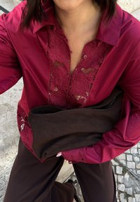 Burgunderfarbene Bluse mit floralen Spitzenakzenten und einer geknöpften Leiste, kombiniert mit einer dunkelbraunen Clutch, getragen über braunen Hosen.