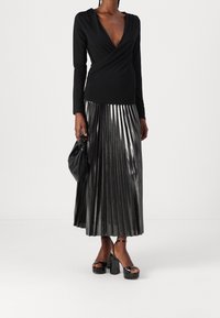 Top nero a maniche lunghe con scollatura a V profonda, abbinato a una gonna plissettata d'argento metallizzato. Sandali neri con tacco e una piccola borsa nera completano il look.