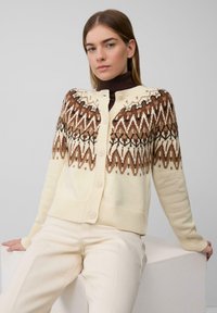 Sweter z dzianiny w kolorze kremowym z brązowym, geometrycznym wzorem na karczku. Posiada guziki z przodu oraz ściągacze przy mankietach i u dołu.