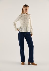 Bijela bluza s dugim rukavima i naboranim teksturom te visokim ovratnikom, uparena s tamnoplavim zvonolikim trapericama i smeđim gležnjačama s potpeticom.