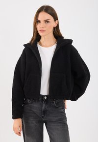 Sort cropped jakke laget av teksturert fleece, med glidelås, hette og framfester, brukt over en hvit skjorte og mørke jeans.