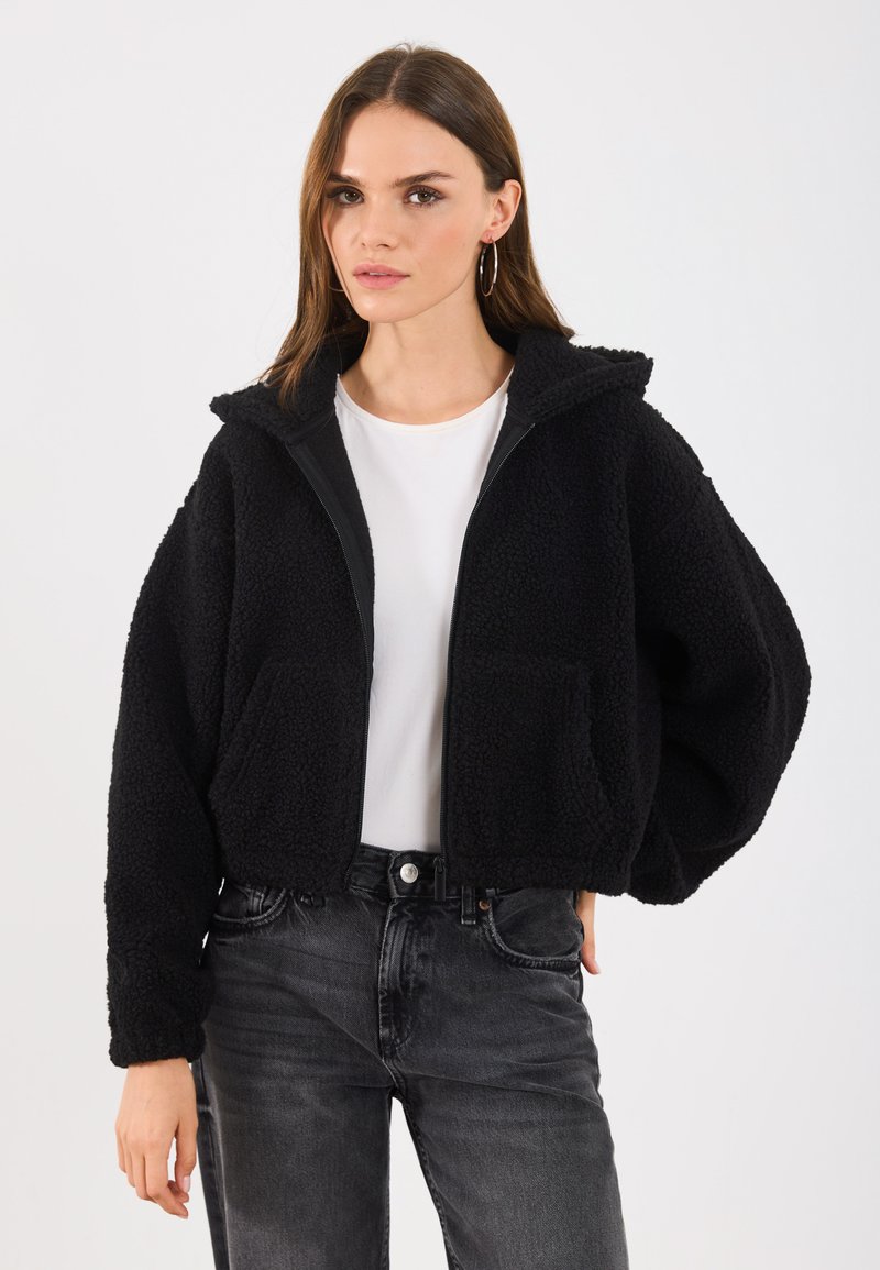 Čierna skrátená bunda z texturovaného fleece, s celoprsťazovým zipsom, kapucňou a prednými vreckami, oblečená na bielej košeli a tmavých džínsoch.