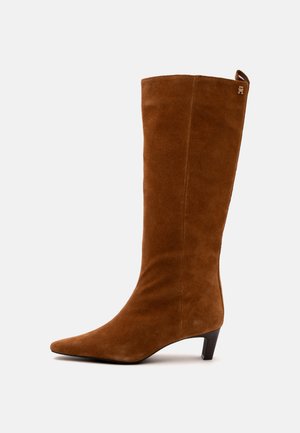 Botte mi-mollet en daim marron avec un bout pointu, un talon bas incliné et un petit logo en métal près du bord supérieur sur fond blanc.