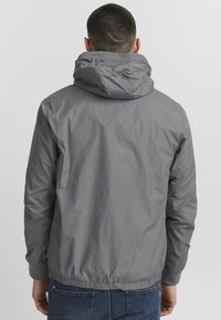 Grå nylonjakke med hætte, elastiske mansjetter og talje. Let glans med minimalistisk design, uden synlige mønstre eller dekorationer.
