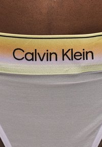 Λάστιχο εσωρούχου Calvin Klein σε κίτρινο βαθμιδωτό χρώμα με μαύρο κείμενο σε φόντο μαυρισμένου δέρματος, με διάφανο λευκό δίχτυ ύφασμα κάτω από το λάστιχο.