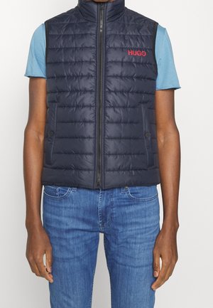 Homme portant une veste matelassée sans manches bleu marine avec un logo rouge "HUGO" sur une chemise bleu clair à manches courtes et un jean bleu.