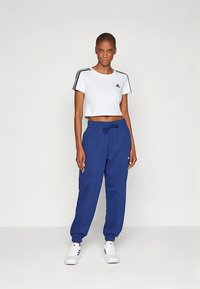 Mulher em pé, vestindo uma t-shirt Adidas branca cropped, calças de treino azuis e ténis Adidas brancos, contra um fundo claro e simples.