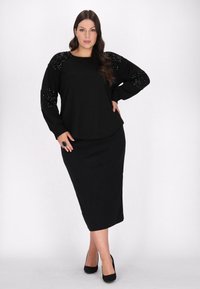 Sort langærmet top med pailletbesatte skuldre, parret med en tætsiddende sort nederdel. Modellen poserer i sorte hæle. Enkel, strømlinet design.