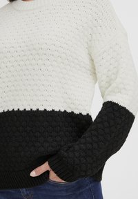 Pulover texturat din tricot, cu o secțiune superioară crem și o secțiune inferioară neagră, având manșete și tiv ondulate pentru accent.
