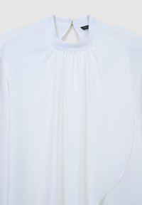 Weiße Bluse mit hochgerundetem Ausschnitt, gerafftem Stoffdetail unter dem Kragen, langen Ärmeln und einer Knopfverschluss hinten.