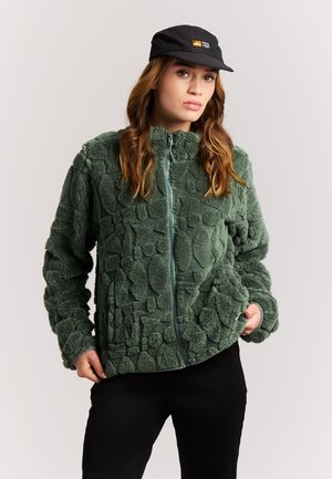 Vrouw draagt een groene, getextureerde fleecejas met rits en zwarte pet met logo, staat met handen in de jaszakken tegen een effen achtergrond.