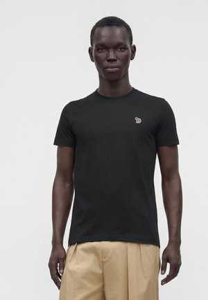 Manden bærer en ensfarvet sort t-shirt med korte ærmer med en lille broderet flerfarvet zebra på brystet og beige bukser.