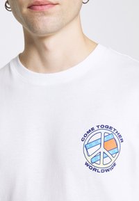 Hvid bomulds T-shirt med et cirkulært fredssymbol grafik med blå og orange accenter og teksten "KOM SAMMEN VERDEN OVER" omkring det.
