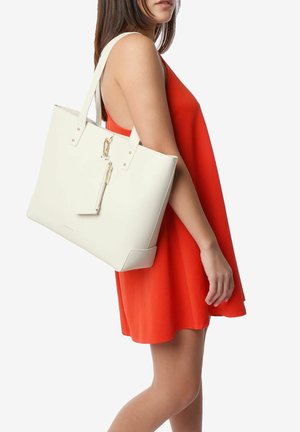 Witte tote bag met een gestructureerd oppervlak, gouden rits en een afneembare tag, vastgehouden door een persoon in een rode jurk. Eenvoudig, modern ontwerp.