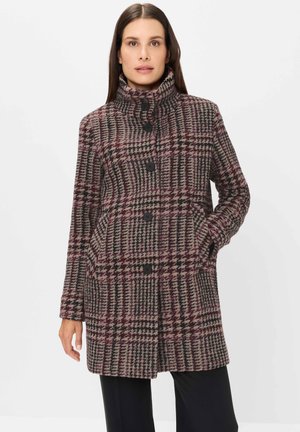 Frau trägt einen knielangen karierten Mantel mit großen schwarzen Knöpfen, hohem Kragen und Seitentaschen, posiert mit einer Hand in der Tasche.