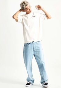 Maglietta bianca di cotone con piccolo logo nero, abbinata a jeans larghi azzurri e sneakers nere e bianche. Outfit semplice e casual.